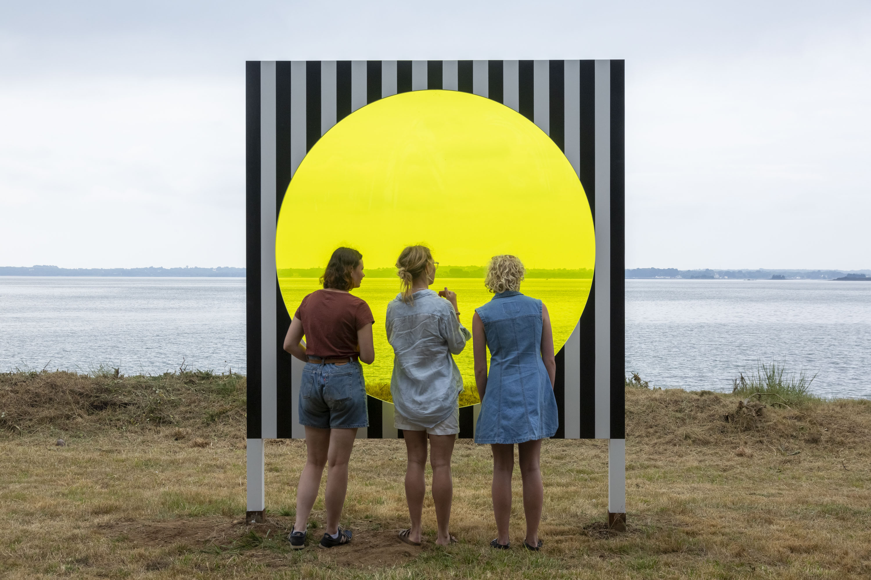 Trois personnes devant une installation présentant un disque jaune transparent devant un paysage de mer.