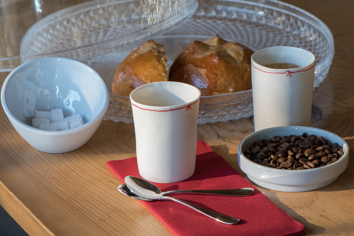 Sur une table en bois, deux tasses en céramique blanche accompagnées d'une coupelle de grains de café, d'un bol de sucre, d'une cuillère et d'un plateau avec petites brioches.