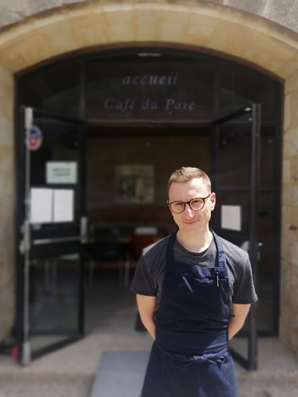 Portrait d'un homme en tablier devant l'entrée d'un café.