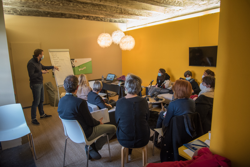 Vue d'un conférencier debout, montrant des écrits sur un paper board, à un groupe d'adultes assis regroupés autour d'une grande table.