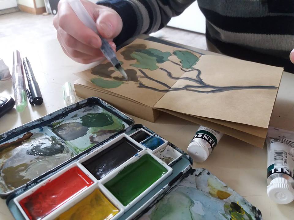 Gros plan sur une main réalisant l'aquarelle d'un arbre sur un carnet, avec du matériel de peinture autour.