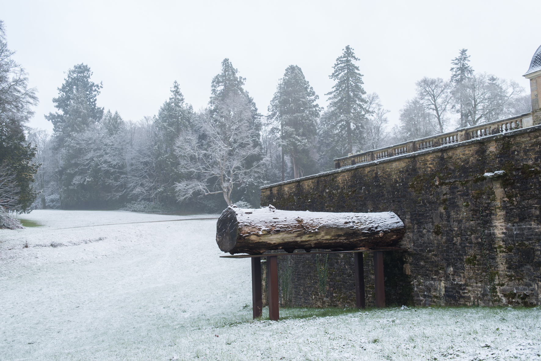 Oeuvre d'art sous la neige exposée contre un mur en pierres avec de grands arbres en arrière plan.