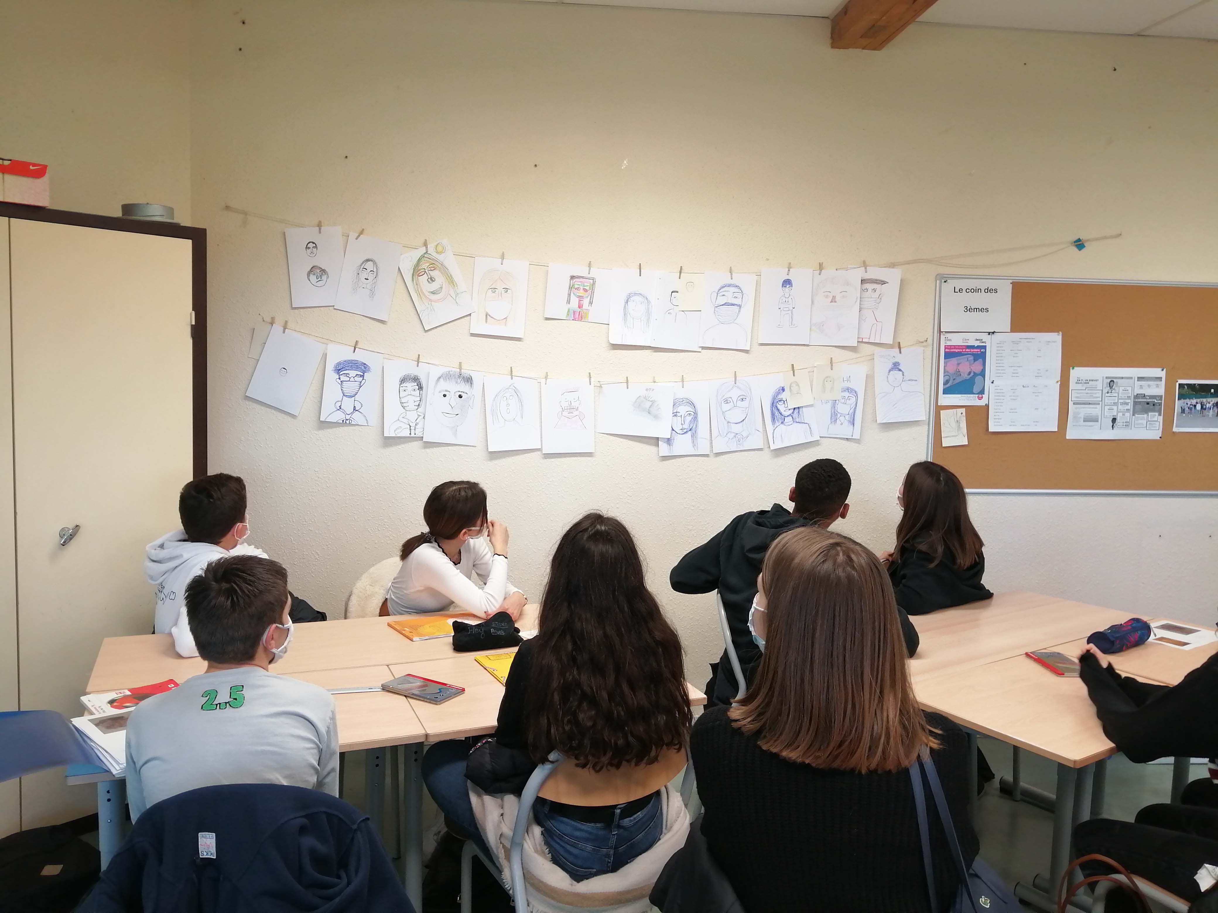 Des élèves assis à leurs tables regardent les autoportraits qu'ils ont réalisés en classe.