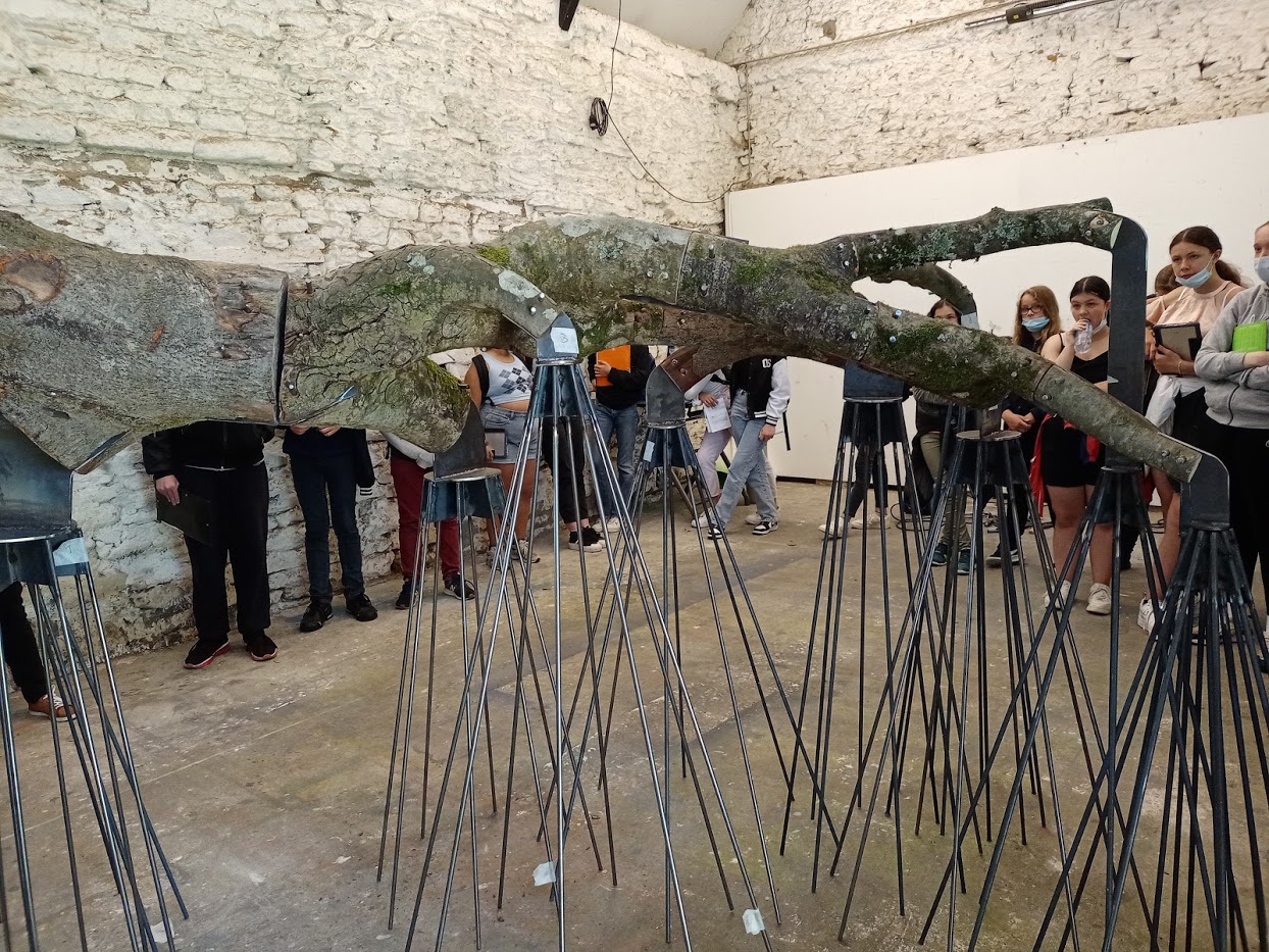 Oeuvre composée d'un tronc reposant sur de multiples pieds en métal. Des élèves sont réunis autour de l'oeuvre dans un atelier.