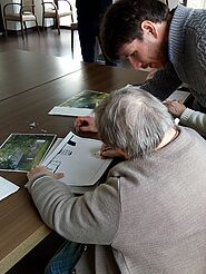 Personne âgée réalisant le dessin d'une maison, assise à une table et aidée par un homme.