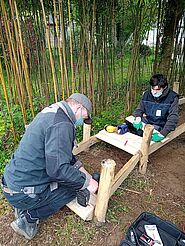 A genoux dans un bois, deux hommes se faisant face clouent des planches en bois sur une structure formant un chemin.