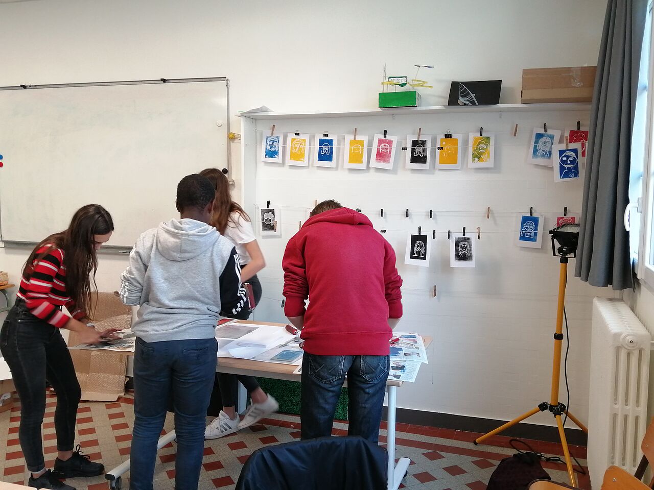 Des élèves réalisent l'impression de leur autoportrait devant un mur où sont accrochés d'autres réalisations en train de sécher.